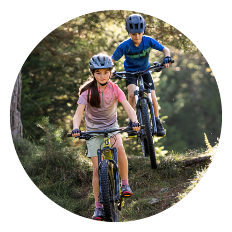 Zwei Kinder mit Helmen fahren Mountainbikes auf einem Waldweg. Das Mädchen vorne trägt ein rosa-grünes T-Shirt, der Junge hinten ein blaues. Sonnenlicht fällt durch die Bäume und hebt die natürliche Umgebung hervor.