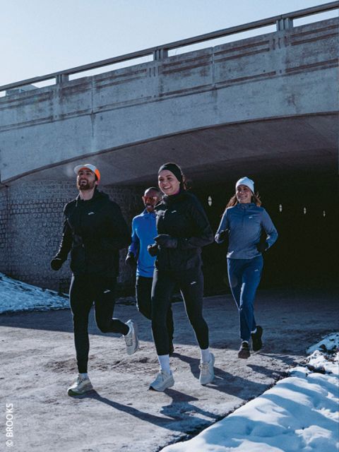 Vier Personen in Winterlaufkleidung joggen gemeinsam auf einem verschneiten Weg unter einer Betonbrücke, lächeln und genießen die Bewegung im Freien.