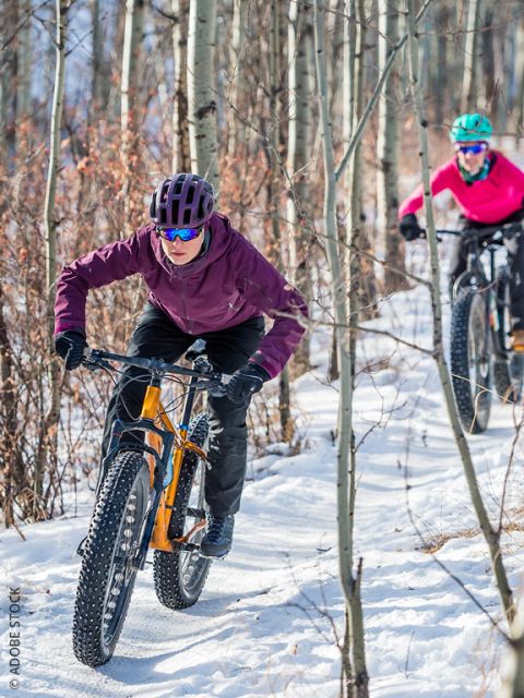 Zwei Radfahrer in Winterkleidung fahren auf einem schmalen, schneebedeckten Weg durch einen Wald mit schlanken Bäumen, durch deren Äste das Sonnenlicht fällt.