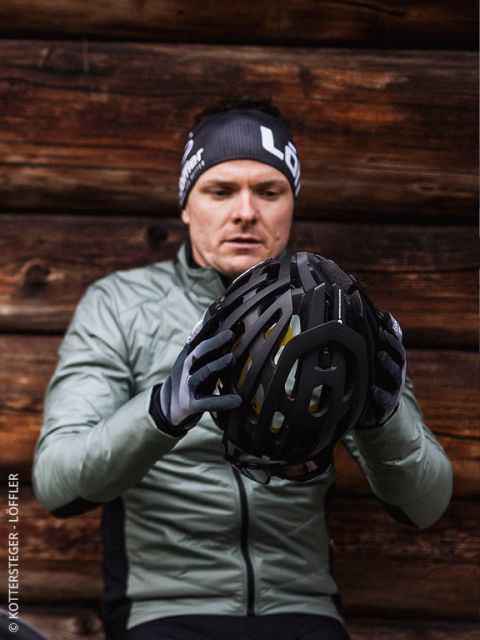 Ein Mann in grüner Jacke und Handschuhen prüft einen schwarzen Fahrradhelm und sitzt vor einer Holzwand. Er trägt ein schwarzes Stirnband mit weißer Schrift.