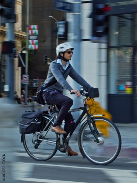 Ein Mann, der einen Helm, eine Sonnenbrille und Geschäftskleidung trägt, fährt mit einem Fahrrad mit Gepäcktaschen durch eine Straße in der Stadt, wobei der unscharfe Hintergrund die Bewegung anzeigt.