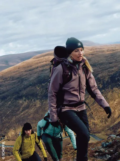 Drei Wanderer mit Outdoor-Ausrüstung und Rucksäcken erklimmen einen felsigen Hang mit braunen, sanften Hügeln im Hintergrund unter einem bewölkten Himmel. Der führende Wanderer trägt eine lila Jacke und eine Mütze.