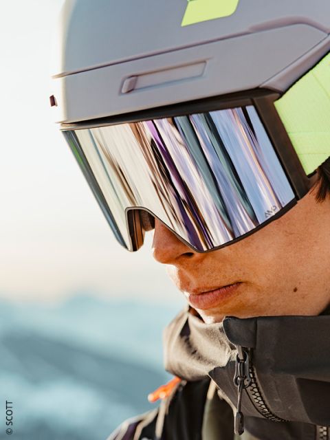 Nahaufnahme einer Person, die einen grauen Skihelm, eine reflektierende Skibrille und eine dunkle Jacke trägt, mit einer verschwommenen Schneelandschaft im Hintergrund.