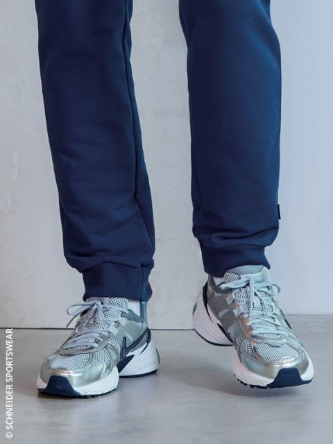 Eine Person in marineblauen Jogginghosen und grauen Laufschuhen mit weißen Sohlen steht in einem Innenraum auf einem hellen Boden.