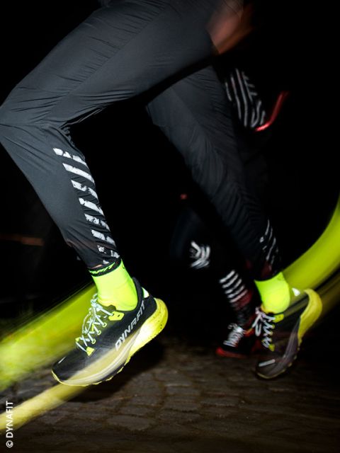 Eine Nahaufnahme von zwei Personen, die nachts laufen. Das Hauptaugenmerk liegt auf ihren Beinen, die schwarze Sporthosen, neongelbe Socken und gelb-schwarze DYNAFIT-Laufschuhe tragen, wobei die Bewegungsunschärfe die Geschwindigkeit betont.