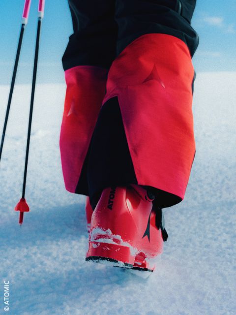 Eine Person in hellbrauner Skihose und grauen Skistiefeln steht mit Skistöcken im Schnee. Die Skistiefel haben rote Akzente und ein Logo mit der Aufschrift "VAN DEER".