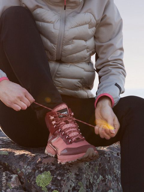 Eine Person in heller Pufferjacke und schwarzer Hose sitzt auf einem Felsen und bindet die Schnürsenkel eines kastanienbraunen Wanderschuhs, während die Abendsonne die Szene sanft beleuchtet.