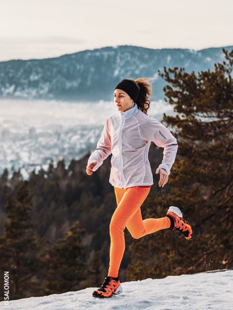 Eine Frau in einer weißen Jacke und leuchtend orangefarbenen Leggings läuft auf einem verschneiten Weg mit Kiefern und Bergen im Hintergrund. Der Himmel ist bewölkt und die Landschaft unter ihr ist teilweise mit Schnee bedeckt.