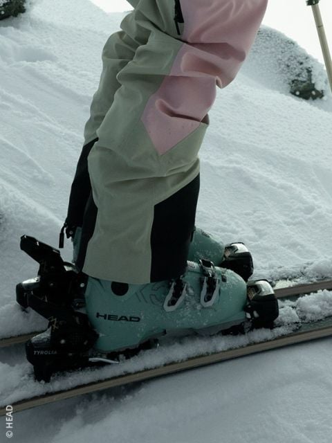 Eine Person in mintgrünen HEAD-Skistiefeln und beige-rosa Skihosen steht auf Skiern im frischen Schnee.