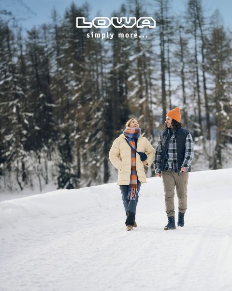 Eine Frau und ein Mann gehen gemeinsam auf einem verschneiten Weg, in Winterkleidung und lächelnd. Schneebedeckte Bäume säumen den Hintergrund. Über ihnen erscheint der Text "LOWA einfach mehr...".