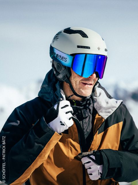 Ein Skifahrer, der einen weißen Helm, eine reflektierende Brille, eine schwarz-braune Jacke und Handschuhe trägt, schließt seine Jacke im Freien mit verschneiten Bergen im Hintergrund ab.