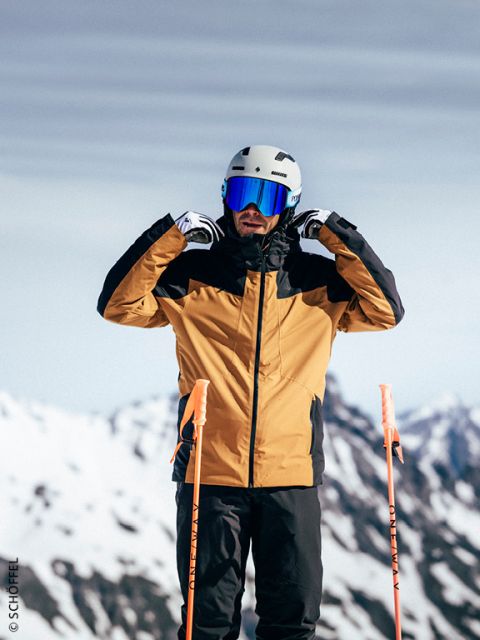 Ein Skifahrer in einer gelb-schwarzen Jacke rückt seinen Helm und seine Skibrille zurecht und steht auf einer verschneiten Piste mit Bergen im Hintergrund.