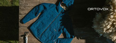 Eine blaue gesteppte Ortovox-Jacke mit Kapuze liegt flach auf einer Holzfläche neben Wolle und Scheren, wobei das Sonnenlicht Schatten wirft.