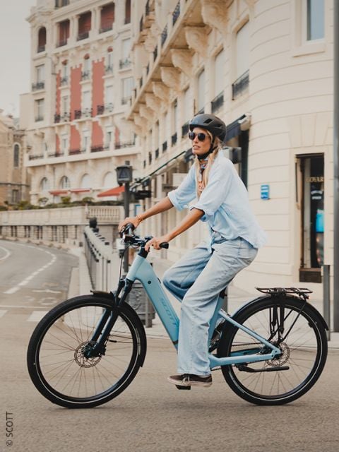 Eine Person, die einen Helm und eine Sonnenbrille trägt, fährt mit einem hellblauen Fahrrad auf einer von historischen Gebäuden gesäumten Stadtstraße.