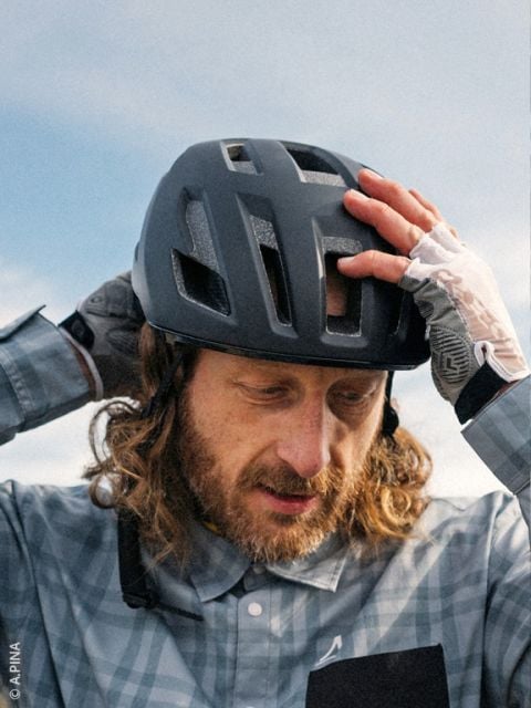 Ein Mann mit langen Haaren und Bart rückt seinen schwarzen Fahrradhelm zurecht, er trägt ein kariertes Hemd und fingerlose Handschuhe, im Hintergrund ist der Himmel zu sehen.