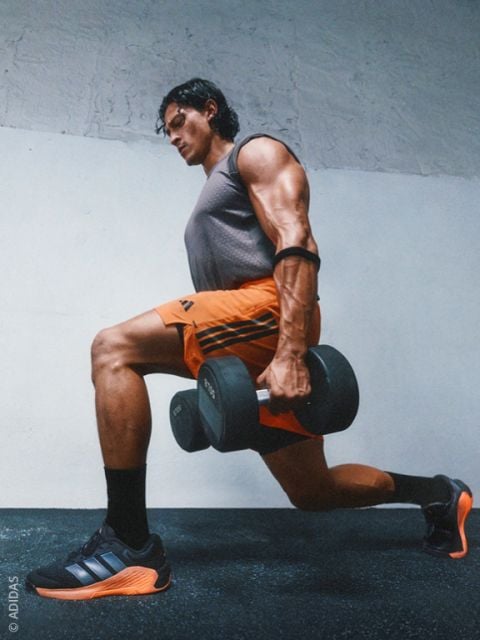 Ein muskulöser Mann in Sportkleidung führt in einem Fitnessstudio einen Ausfallschritt mit Hanteln aus und trägt schwarze und orangefarbene Adidas-Turnschuhe.