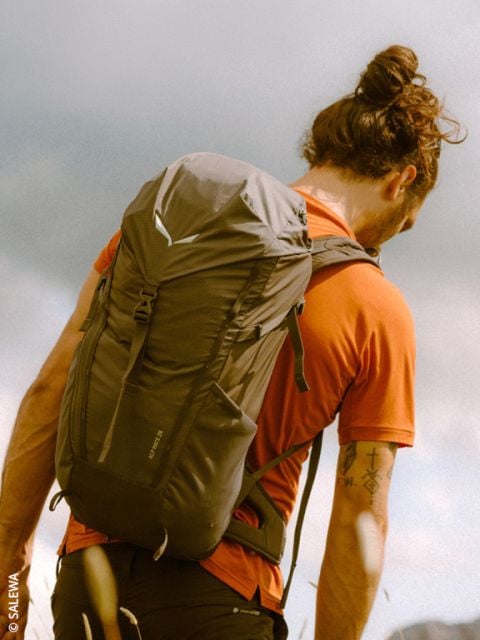 Eine Person mit einem Haarknoten, einem orangefarbenen Hemd und einem Rucksack steht im Freien unter einem bewölkten Himmel und blickt in die Ferne.