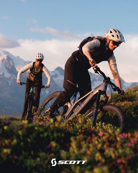 Zwei Radfahrer mit Helmen fahren Mountainbike auf einem malerischen Weg mit Bergen im Hintergrund bei Sonnenuntergang. Das Scott-Logo befindet sich am unteren Rand.
