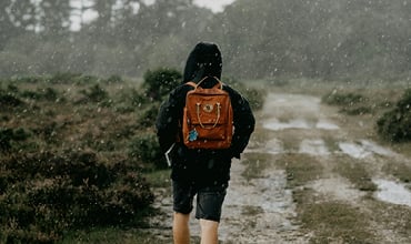 Eine Person mit einem braunen Rucksack geht im Regen auf einem schlammigen Weg, umgeben von Grün und Bäumen.