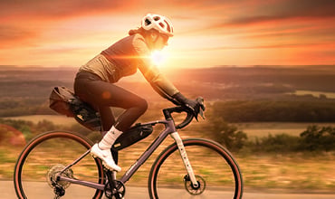Ein Radfahrer fährt bei Sonnenuntergang mit einem Rennrad bergauf, während die Sonne hinter ihm strahlt und im Hintergrund malerische Hügel zu sehen sind.