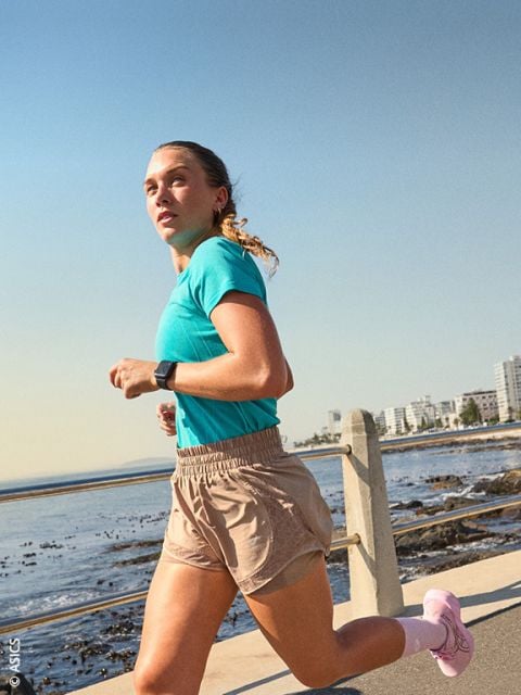 Eine Frau in Sportkleidung joggt an einem sonnigen Tag am Meer entlang, im Hintergrund sind die Gebäude der Stadt zu sehen.